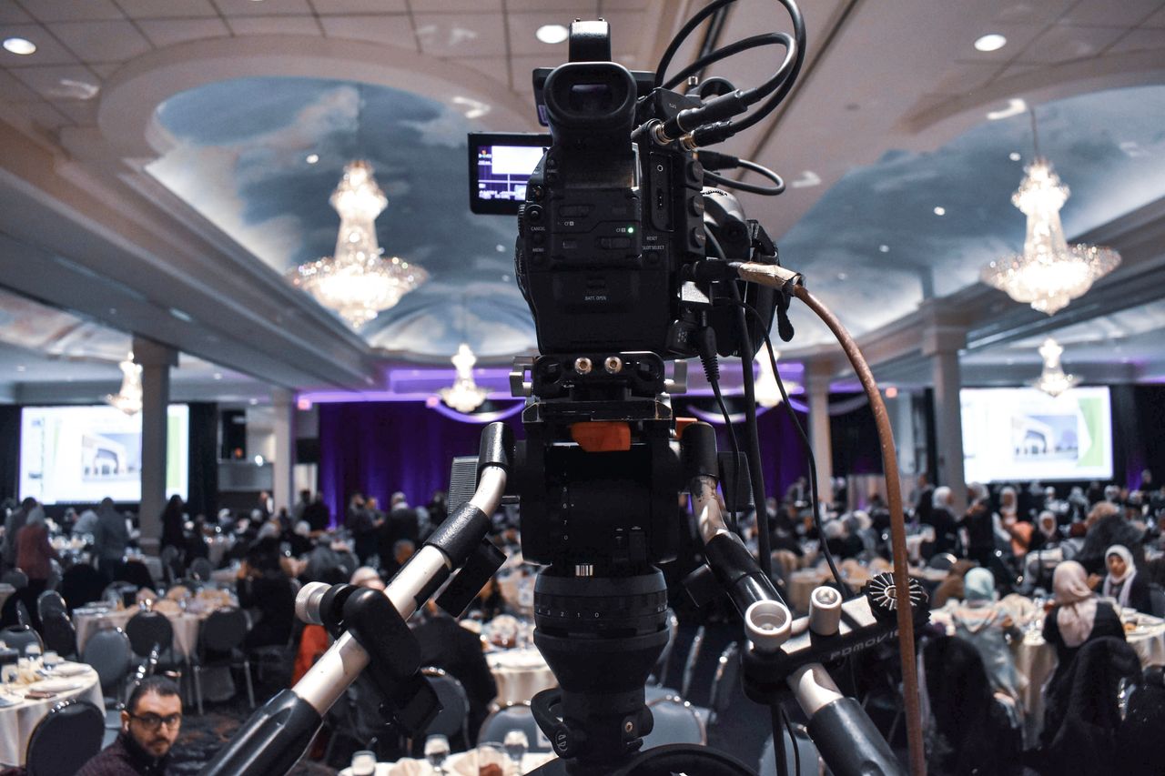 Modern professional video camera placed on tripod filming crowded ballroom during diner party